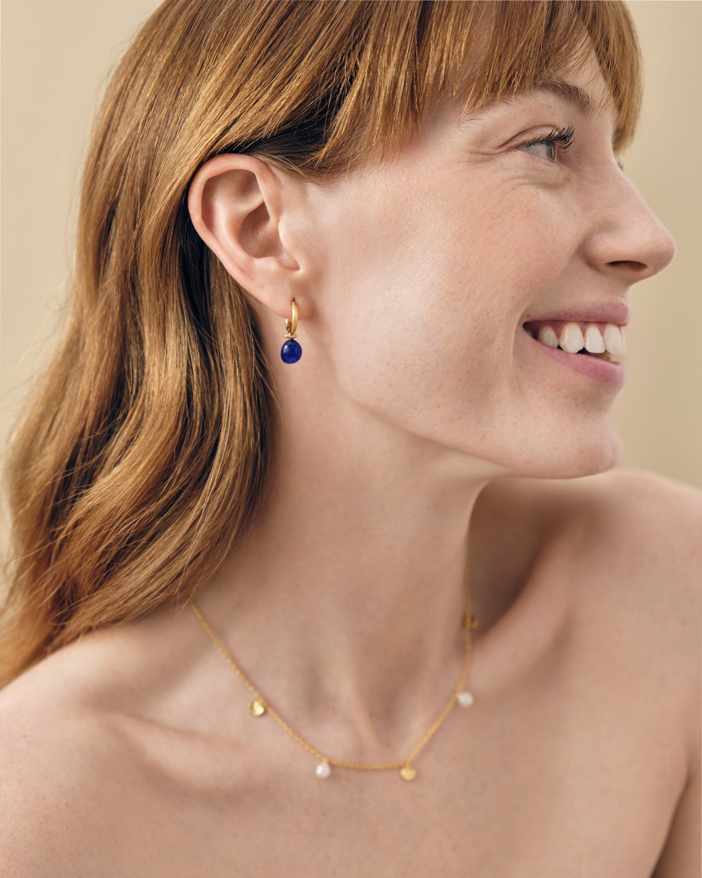 Close-up of a smiling woman wearing Coastal Blue Quartz Earrings, gold-plated hoop style with deep blue quartz stones, paired with a delicate gold necklace, showcasing affordable luxury and ethical craftsmanship.