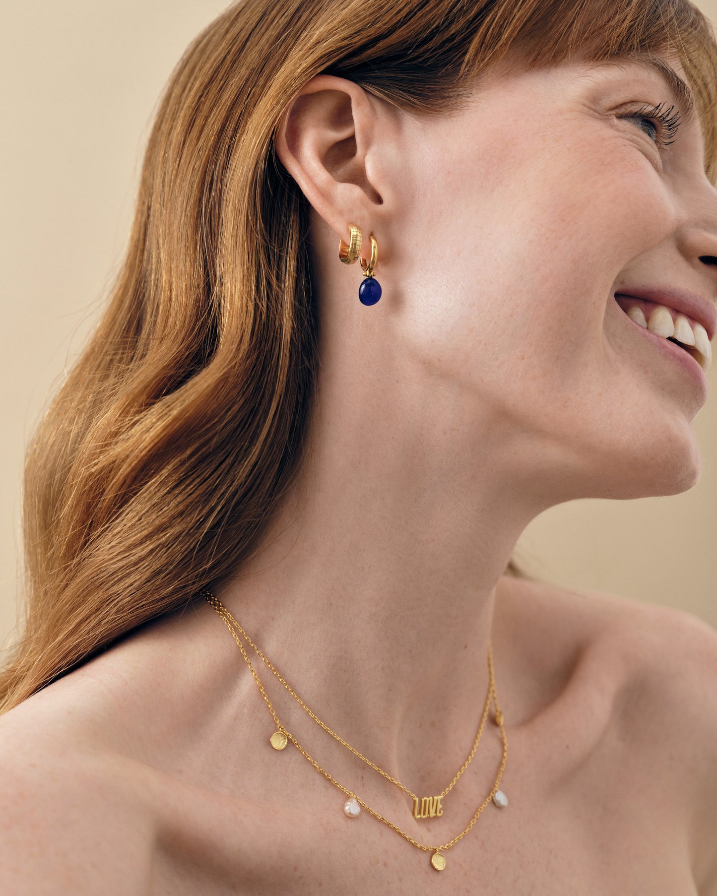 Close-up of a smiling woman wearing Coastal Blue Quartz Earrings, featuring gold-plated sterling silver hoop design with a blue quartz charm, paired with delicate layered gold necklaces.