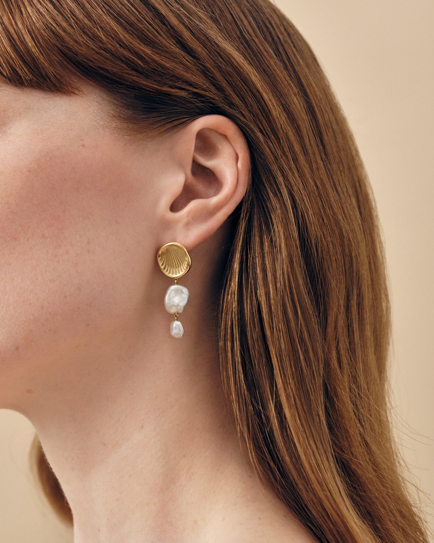 Close-up of a woman's ear wearing Coastal Pearl Statement Earrings featuring gold-plated shell design stud with two dangling pearls, crafted in sterling silver and recycled metals.