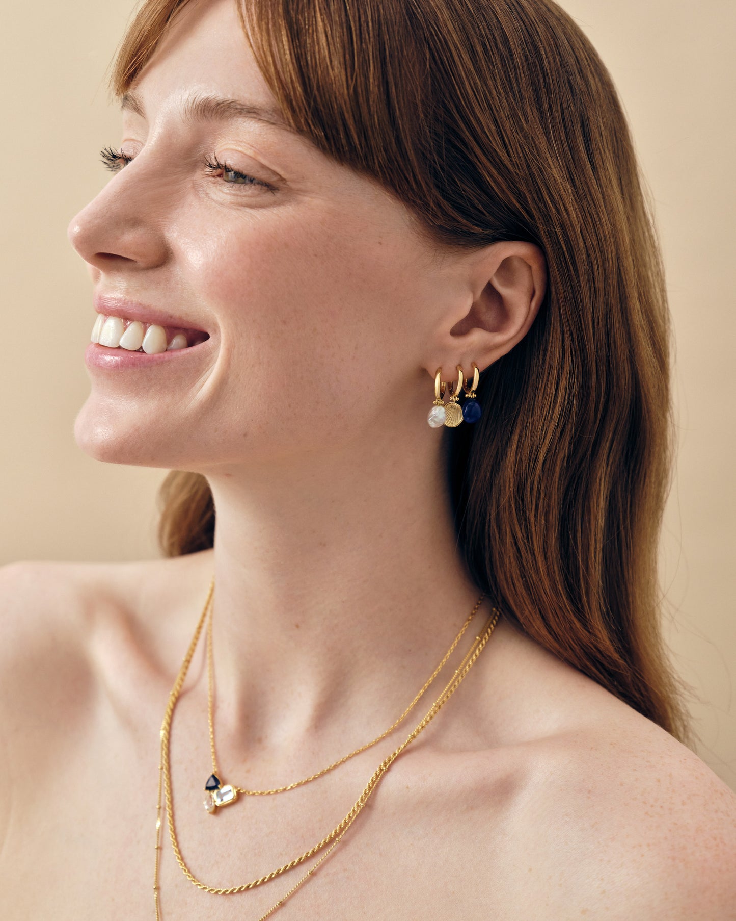 Woman smiling wearing Coastal Shell Earrings in gold-plated and sterling silver stud design, paired with layered gold necklaces, embodying considered jewellery made in recycled metals.