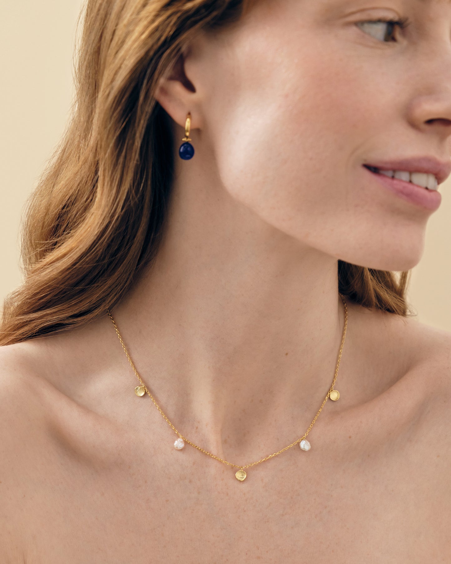 Woman wearing a Coastal Shell and Pearl Station Necklace featuring gold-plated chain with alternating shell and pearl pendants, styled with a gold-plated hoop earring, showcasing affordable, ethically sourced fine jewellery made from recycled metals.