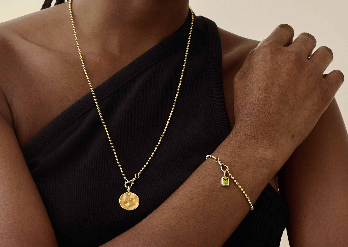 A person wearing a delicate solid gold chain necklace with a round textured pendant and a matching gold bracelet featuring a small charm, styled with a black sleeveless top.
