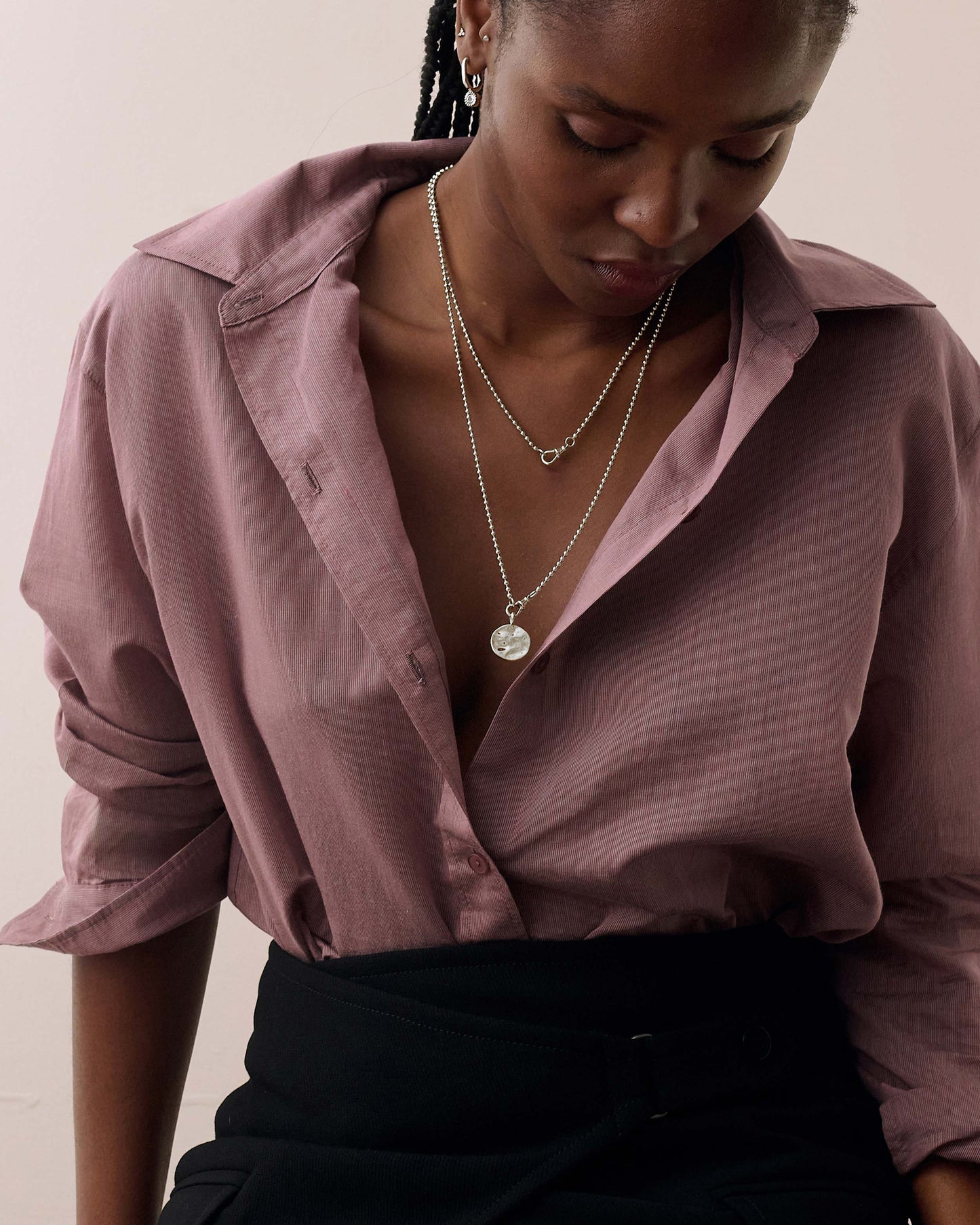 Woman wearing the Coin Globe Necklace - Silver with a pendant and chain in sterling silver, styled with a relaxed mauve shirt and black skirt, showcasing considered, sustainable fine jewellery.