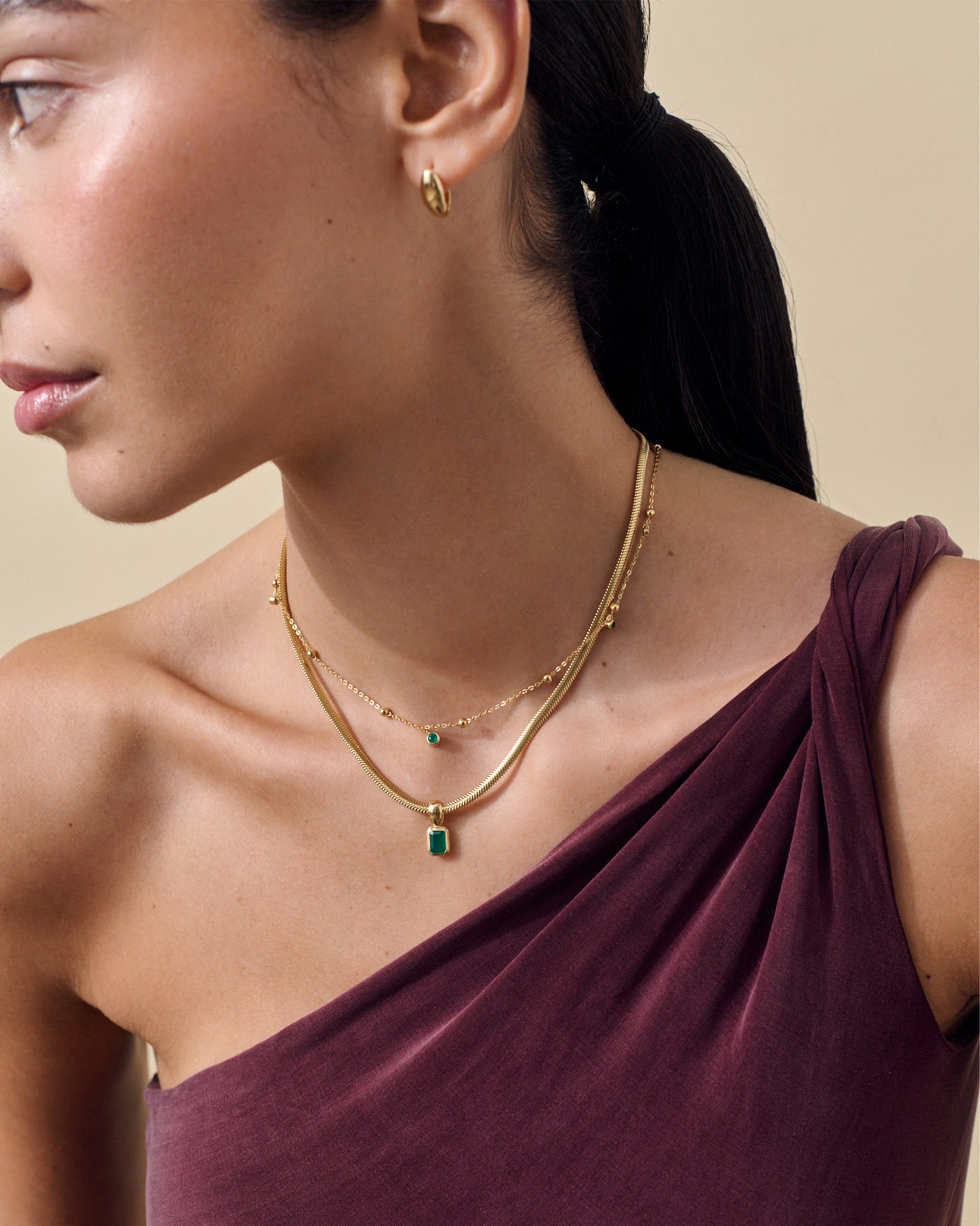 Model wearing layered gold-plated necklaces including the Pebble Green Onyx Necklace pendant, styled with a gold hoop earring and a wine-colored one-shoulder top, showcasing fine jewellery crafted from recycled metals.