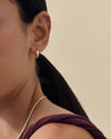Close-up of a woman wearing the Pebble Hoop Earring - Gold, a sleek gold-plated dangle earring paired with a delicate gold necklace, showcasing sustainable fine jewellery crafted from recycled metals and solid craftsmanship.