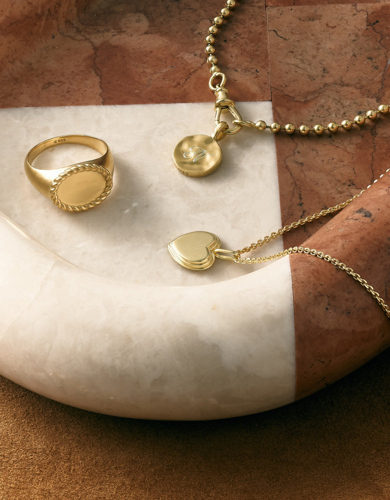 Three solid 14ct gold pieces including a signet ring with a braided edge, a heart-shaped pendant necklace, and a round pendant on a beaded chain, displayed on a curved marble surface.