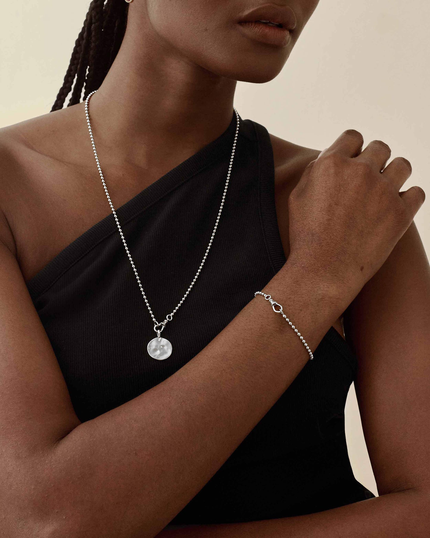 Woman wearing a Coin Globe Necklace - Silver pendant on a sterling silver chain paired with a delicate matching silver bracelet, styled with a black one-shoulder top, showcasing considered jewellery made from recycled metals and crafted to last.