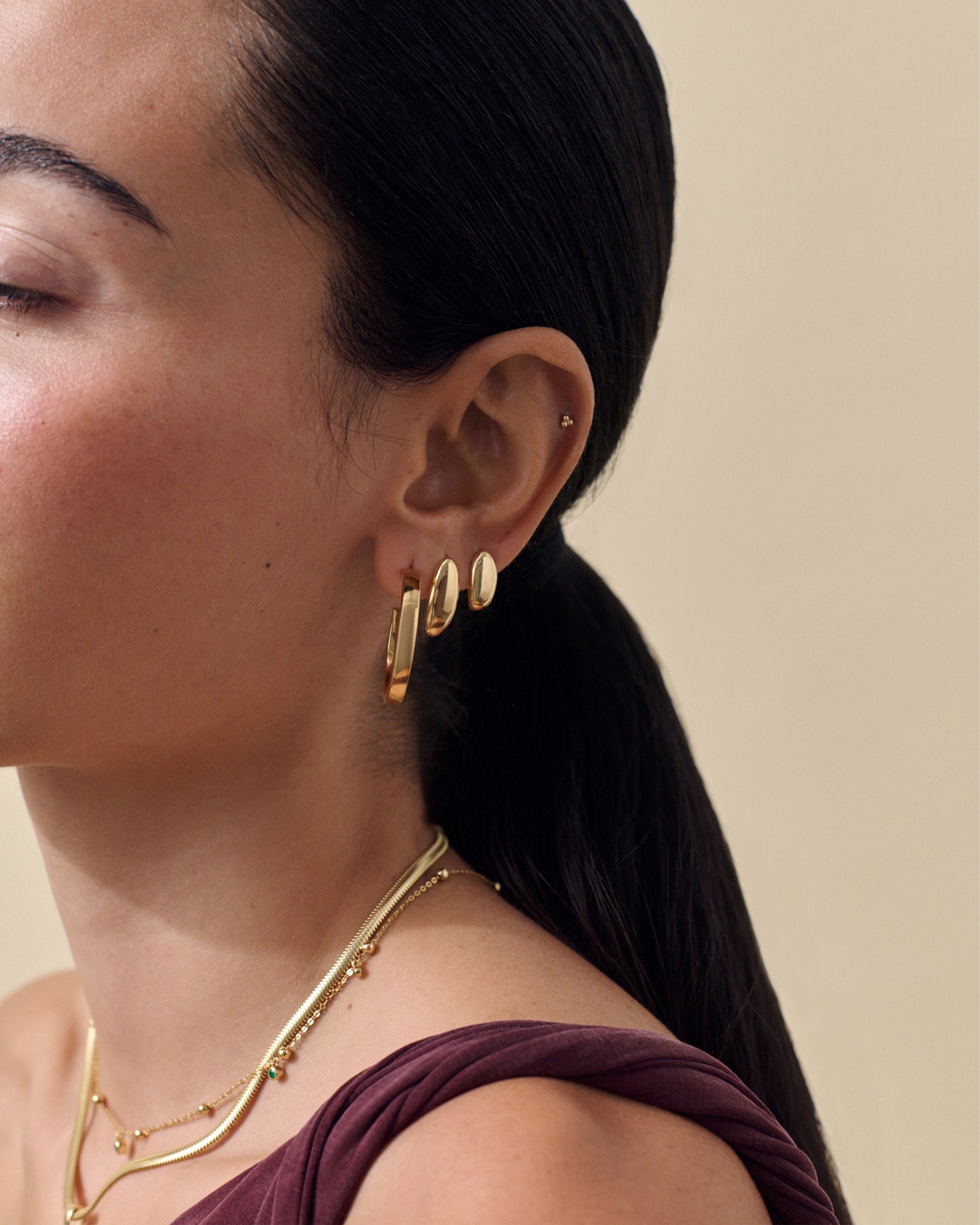Close-up of a woman's ear wearing the Pebble Hoop Earring - Gold along with other gold-plated dangle earrings, showcasing considered jewellery made from recycled metals and solid gold for everyday fine jewellery.