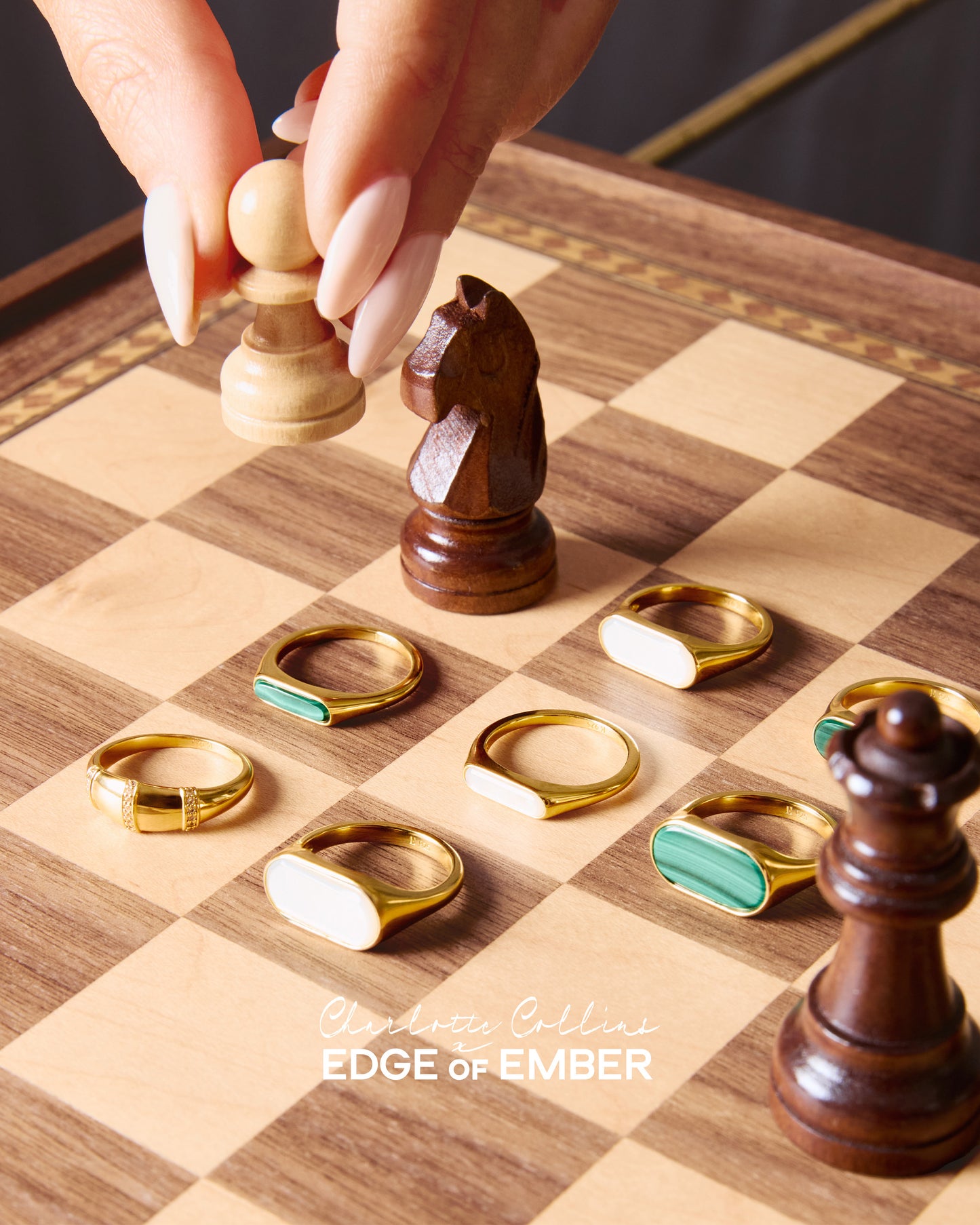 Close-up of a hand moving a chess pawn on a wooden chessboard surrounded by elegant gold-plated rings featuring mother of pearl and green stone inlays, showcasing the Charlotte Collins Mother of Pearl Cocktail Ring.