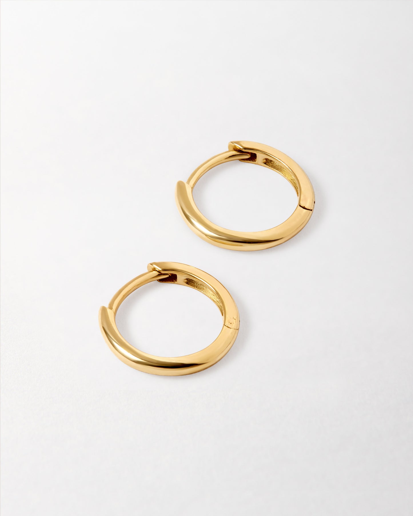 Pair of polished Solid Gold Mini Huggie Hoop Earrings displayed on a light surface, crafted from recycled metals for everyday fine jewellery with sustainable luxury.
