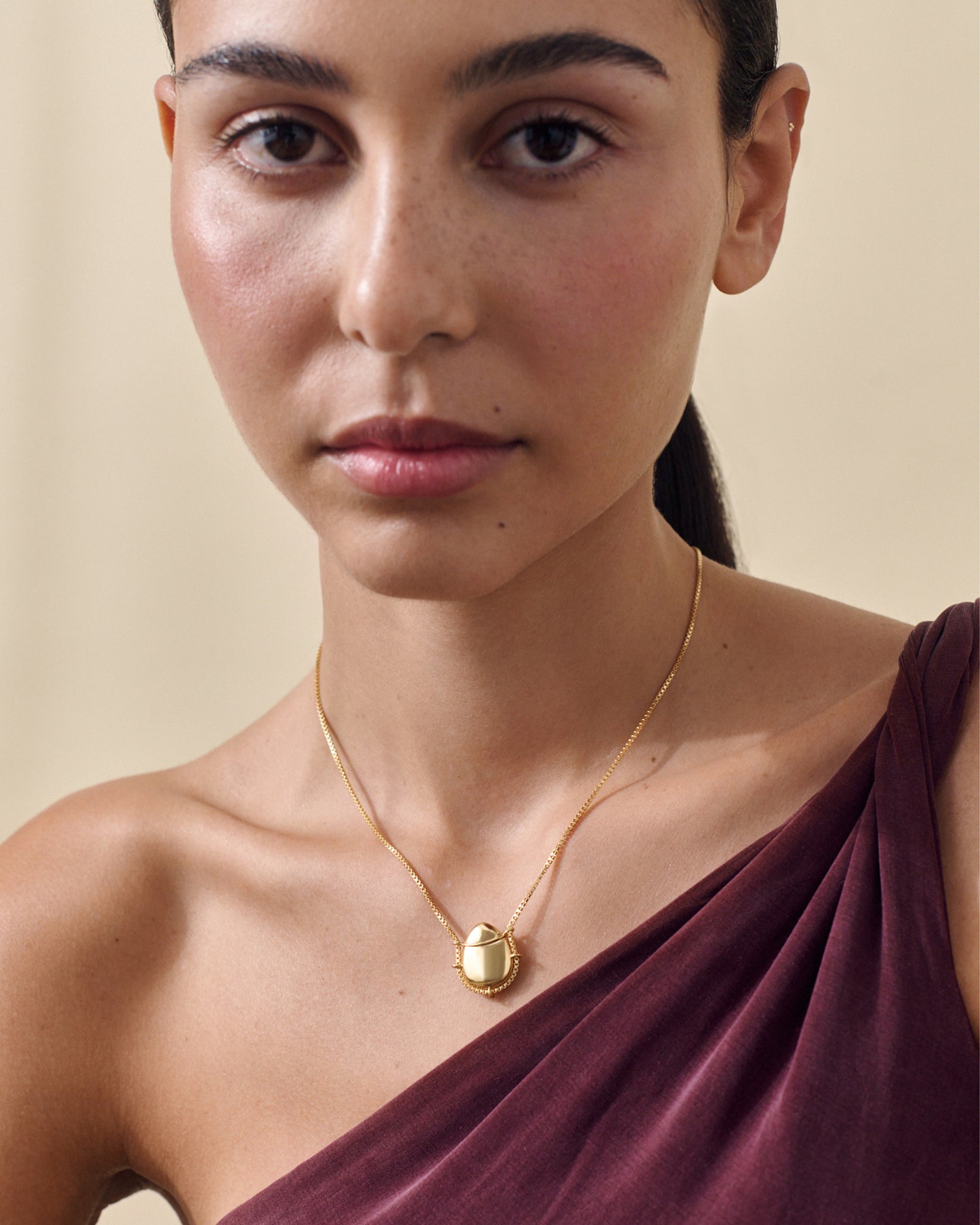 Close-up of a woman wearing a gold Secret Message Locket Necklace with a smooth pendant and fine chain, styled with an elegant off-shoulder burgundy top, showcasing considered jewellery made in recycled metals.