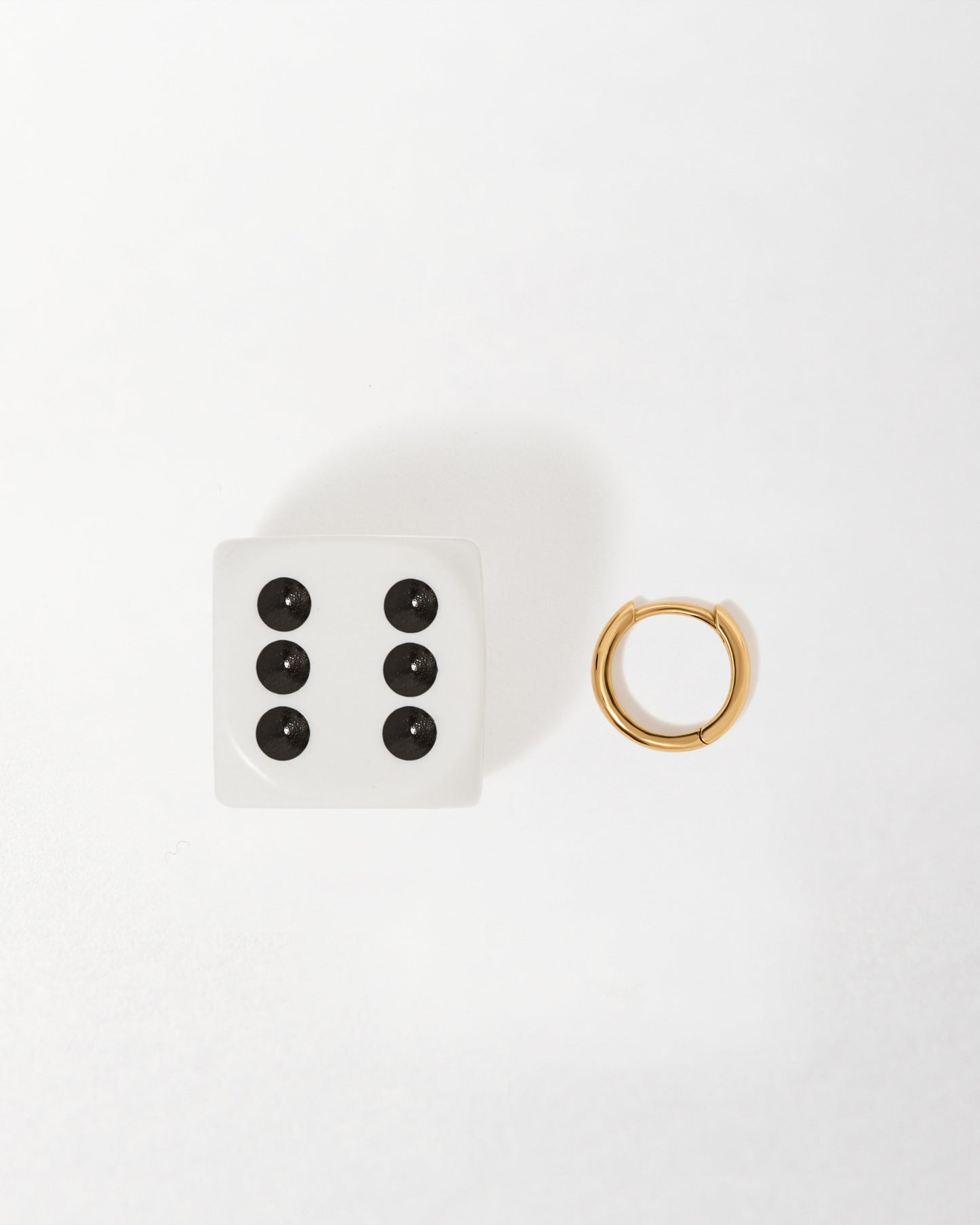 Solid Gold Mini Huggie Hoop Earrings displayed next to a white die, showcasing considered jewellery crafted from recycled metals with solid 14ct gold, built to last.