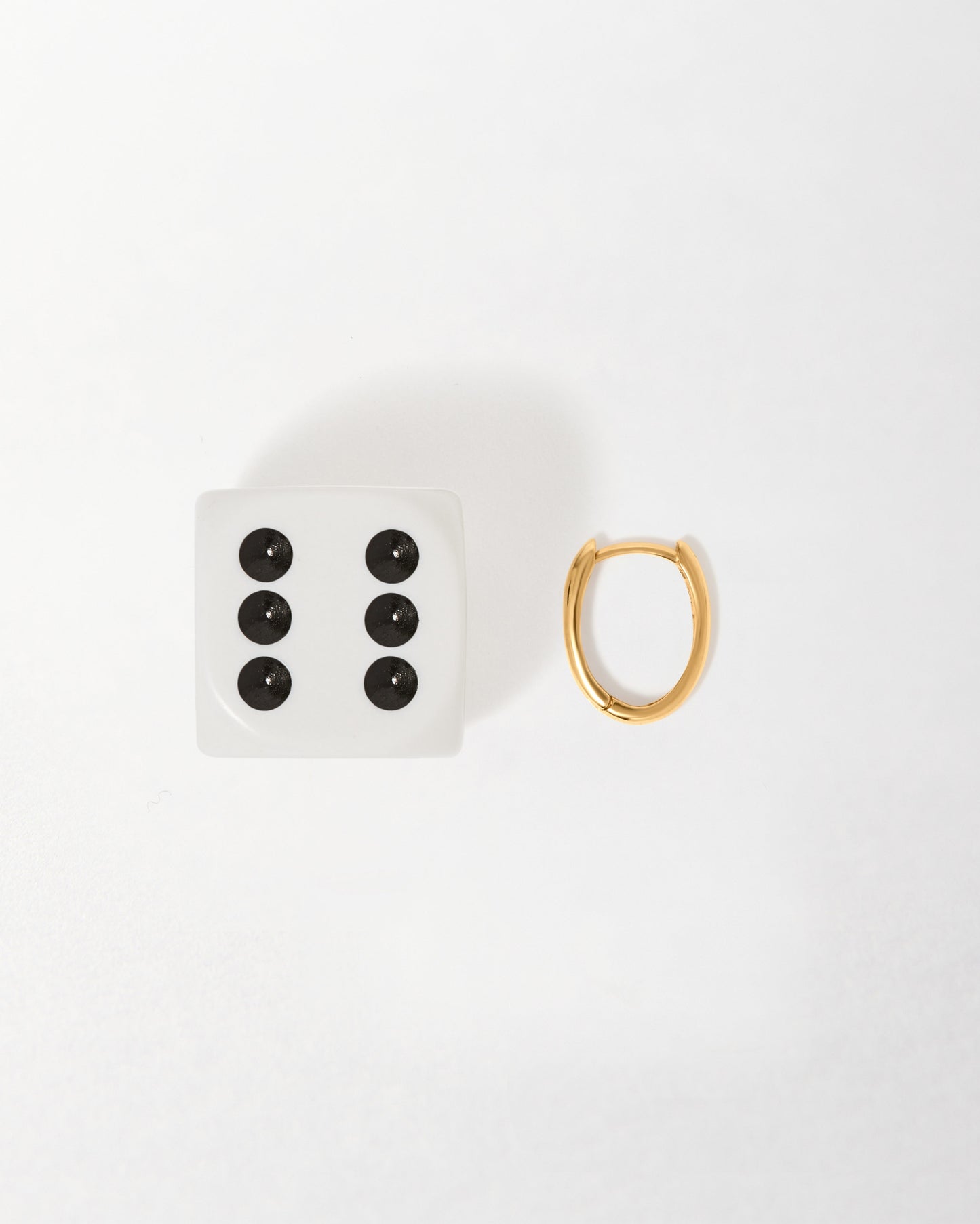 Solid Gold Oval Hoop Earrings next to a die showing six, highlighting the small, elegant 14ct gold huggie design crafted from recycled metals for everyday fine jewellery.