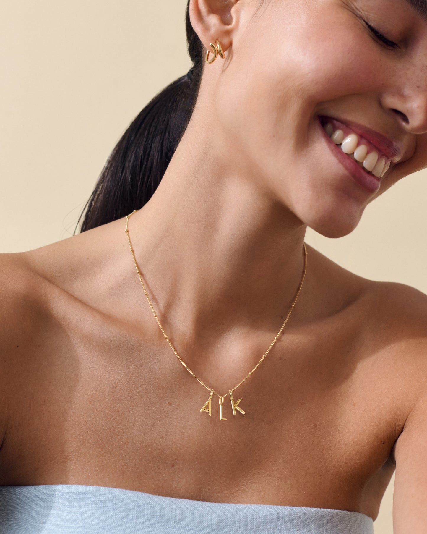Close-up of a smiling woman wearing the Triple Mini Initial Necklace - Gold, featuring delicate gold-plated chain and personalized letter pendants, crafted from recycled metals.