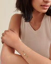 A woman wearing three delicate gold-plated gemstone bracelets stacked on her wrist, showcasing the Deco Opal October Birthstone Bracelet with elegant, sustainable fine jewellery crafted from recycled metals.